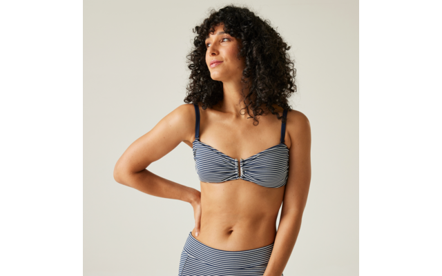 Person mit lockigen dunklen Haaren in blau-weiß gestreiftem Bikinioberteil und hoch sitzendem Bikinihöschen, eine Hand in die Hüfte gestützt, Blick nach links vor einfarbig hellem Hintergrund