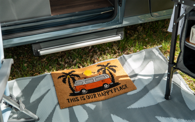 Paillasson avec illustration d'un minibus orange, palmiers, soleil et le texte "THIS IS OUR HAPPY PLACE"; posé sur un tapis d'extérieur gris devant une porte de véhicule ouverte, herbe et pieds de mobilier visibles