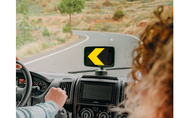 Blick aus dem Fahrzeuginnenraum auf die Hand des Fahrers am Lenkrad, einen Handyhalter mit gelbem Links-Pfeil auf dem Display, eine kurvige Straße voraus und eine Person mit lockigen Haaren vorne rechts