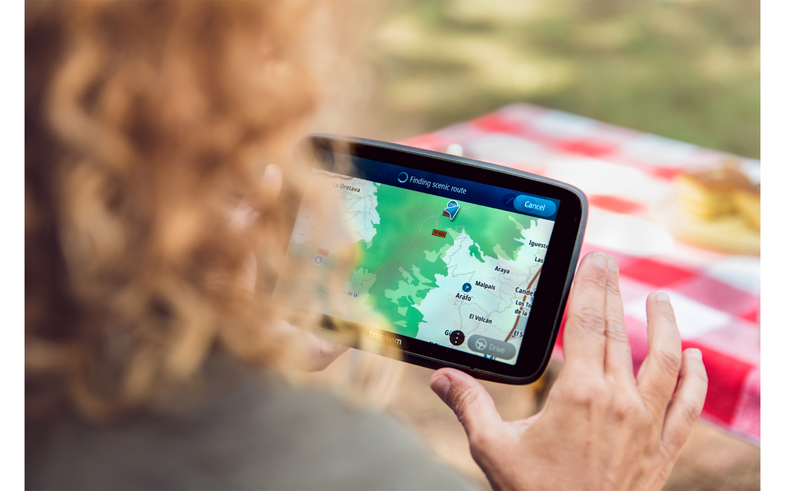 Person von hinten hält ein Tablet, auf dem eine Karten-App mit Route und Markern zu sehen ist; im Vordergrund ein rot-weiß kariertes Picknicktuch