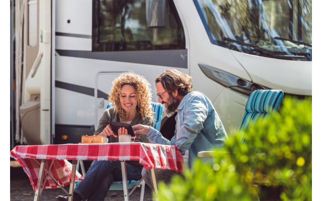 Zwei Erwachsene sitzen an einem kleinen Tisch mit rot-weiß kariertem Tischtuch vor einem weißen Wohnmobil und schauen auf ein Tablet; Tassen auf dem Tisch, eine Person trägt eine Sonnenbrille auf dem Kopf, im Vordergrund ein grüner Strauch