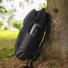 Schwarzer Rucksack lehnt an einem Baumstamm auf Gras; schwarze Thermosflasche in der Seitentasche