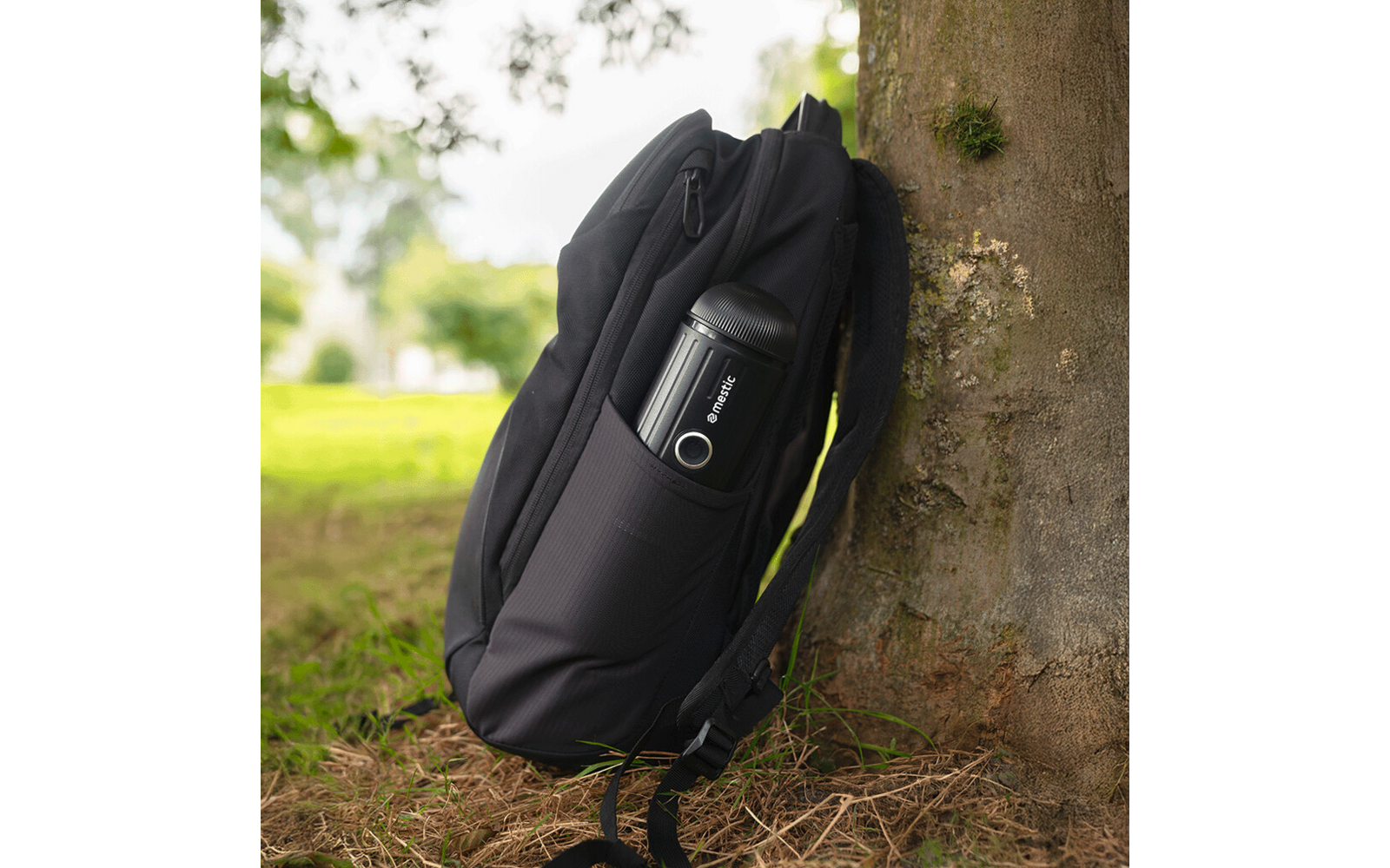 Schwarzer Rucksack lehnt an einem Baumstamm auf Gras; schwarze Thermosflasche in der Seitentasche