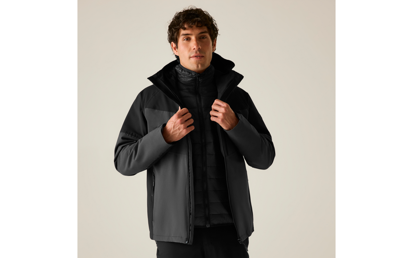 Person mit dunkler Softshelljacke und gesteppter Innenjacke, Hände am Reißverschluss, heller einfarbiger Hintergrund