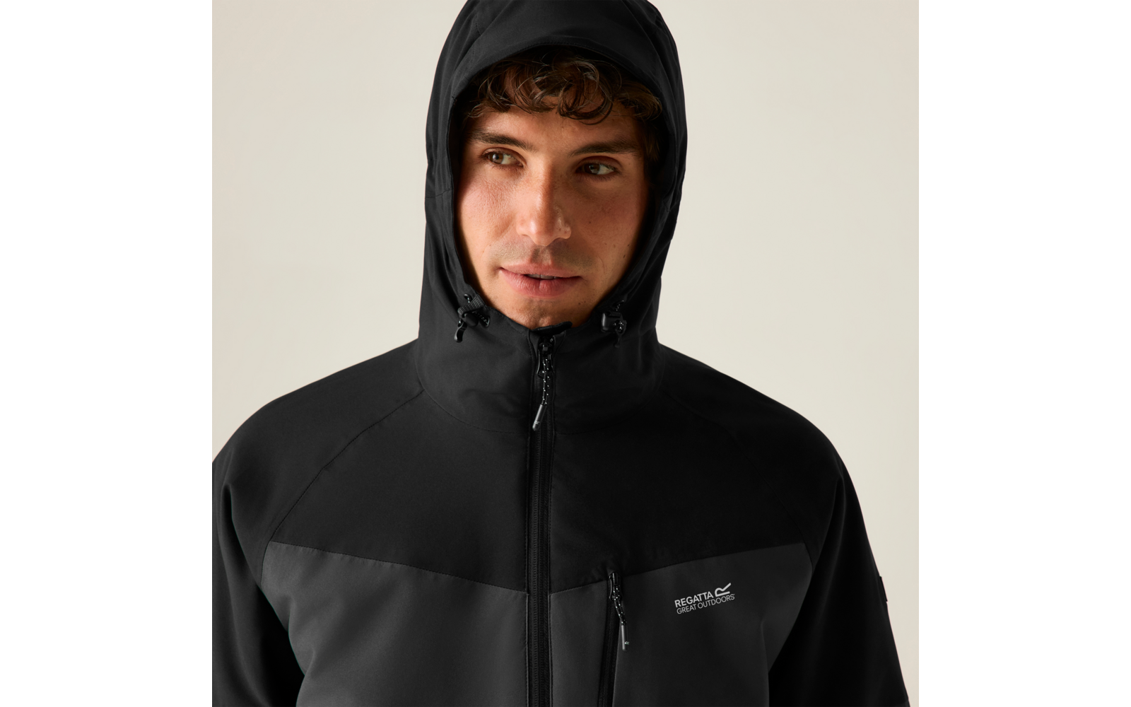 Person mit schwarzer Kapuzenjacke, Reißverschluss bis zum Kinn, sichtbares Logo auf der Brust, Blick nach rechts, heller Hintergrund