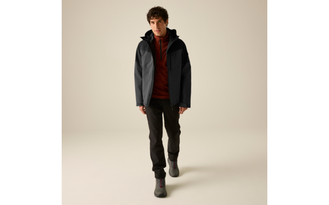 Ein Mann mit lockigem Haar geht vor einem neutralen Hintergrund. Er trägt eine zweifarbige dunkle Jacke, einen rostfarbenen Zip-Pullover, dunkle Hosen und graue Wanderschuhe
