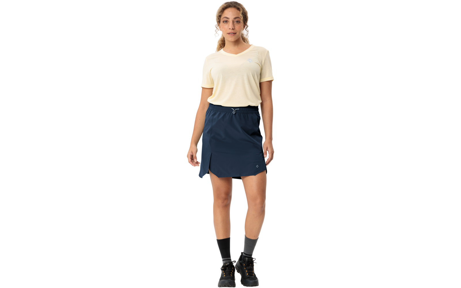 Personne debout de face, porte un t-shirt jaune pâle à manches courtes, une jupe bleu marine avec cordon, des chaussettes noires et des chaussures de randonnée
