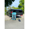 Station wagon scuro con bagagliaio aperto su un'area di parcheggio lastricata; davanti una grande borsa turchese e nera con spallacci e una pagaia nera appoggiata, targa visibile; alberi sullo sfondo