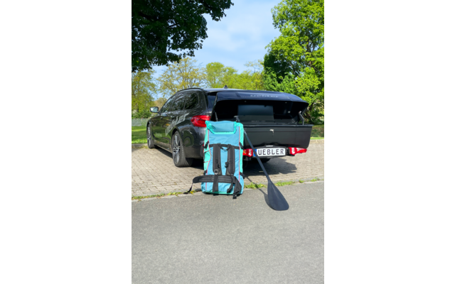 Station wagon scuro con bagagliaio aperto su un'area di parcheggio lastricata; davanti una grande borsa turchese e nera con spallacci e una pagaia nera appoggiata, targa visibile; alberi sullo sfondo