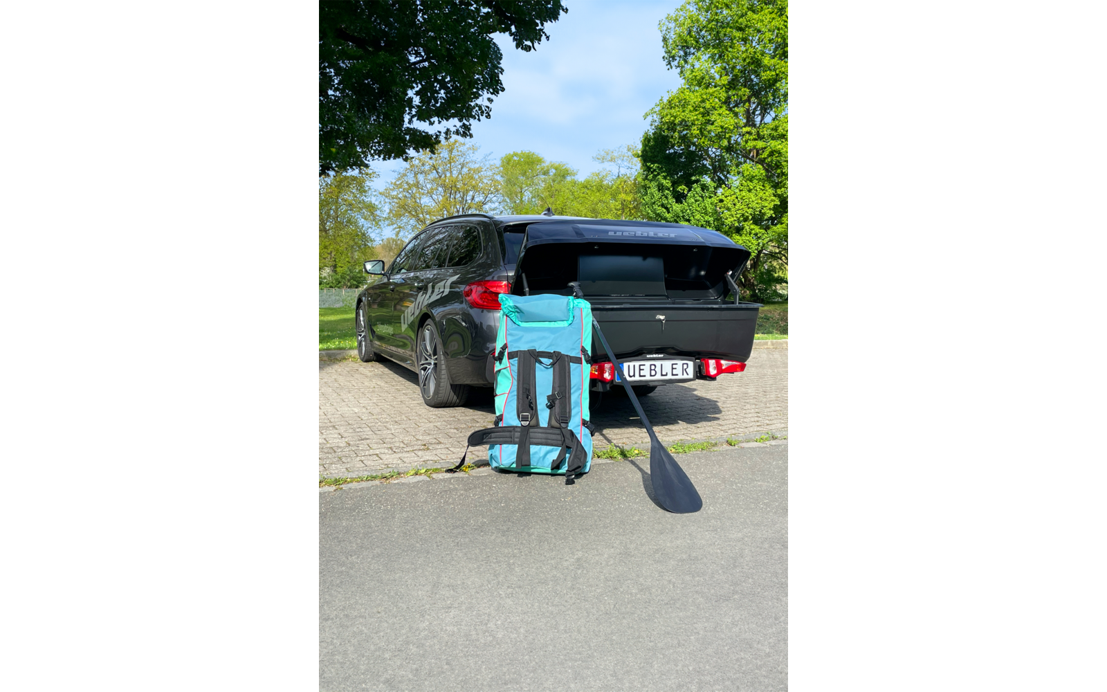 Station wagon scuro con bagagliaio aperto su un'area di parcheggio lastricata; davanti una grande borsa turchese e nera con spallacci e una pagaia nera appoggiata, targa visibile; alberi sullo sfondo