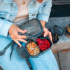 Mains tenant une boîte-repas grise à deux compartiments ; à gauche muesli avec noix et graines, à droite pâte rouge ; personne portant une veste en jean bleu clair et un jean ; à côté un petit bol de raisins et un thermos