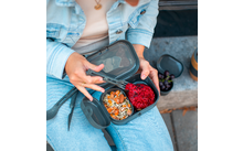 Mains tenant une boîte-repas grise à deux compartiments ; à gauche muesli avec noix et graines, à droite pâte rouge ; personne portant une veste en jean bleu clair et un jean ; à côté un petit bol de raisins et un thermos