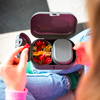 Boîte repas ouverte de couleur violet foncé avec des oursons en gélatine colorés et un récipient gris ; une main tient un ourson rouge ; personne assise jambes croisées en jean bleu et pull rose