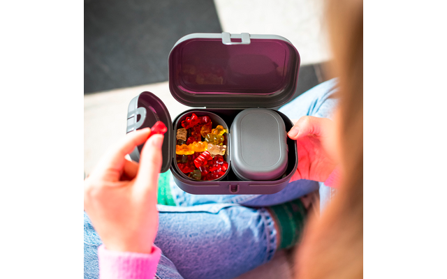Boîte repas ouverte de couleur violet foncé avec des oursons en gélatine colorés et un récipient gris ; une main tient un ourson rouge ; personne assise jambes croisées en jean bleu et pull rose