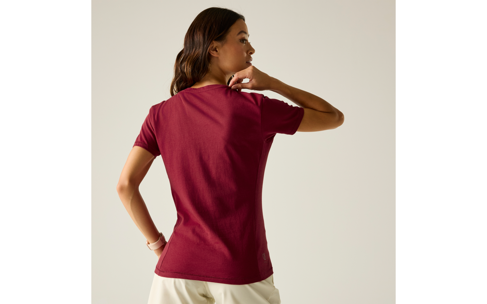 Person von hinten, trägt kurzärmeliges weinrotes T-Shirt und helle Hose, rechte Hand am Nacken, linke Hand an der Hüfte, heller einfarbiger Hintergrund