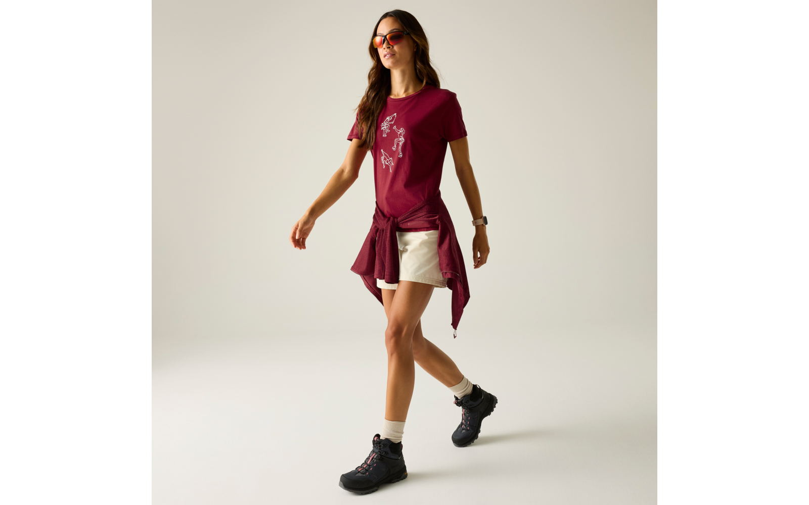 Person geht, trägt burgunderrotes T-Shirt mit weißem Muster, beige Shorts, burgunderfarbenen Pullover um die Hüfte gebunden, schwarze Wanderschuhe, weiße Socken und orangefarbene Sonnenbrille