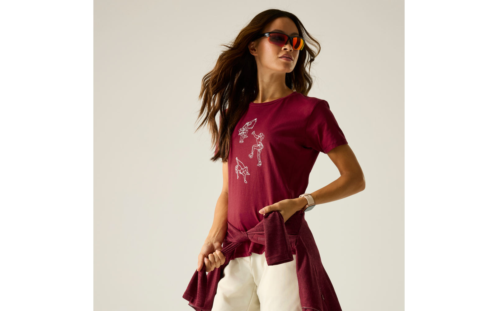 Frau mit Sonnenbrille, bordeauxrotes T-Shirt mit weißen Zeichnungen, Pullover um die Hüfte, weiße Hose, Hand in der Tasche, heller Hintergrund