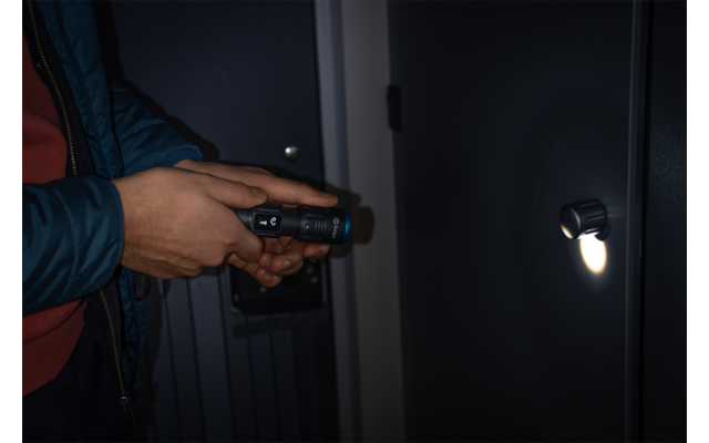 Hände einer Person in blauer Jacke und rotem Pullover halten eine Taschenlampe, deren Licht auf ein rundes Türschloss scheint