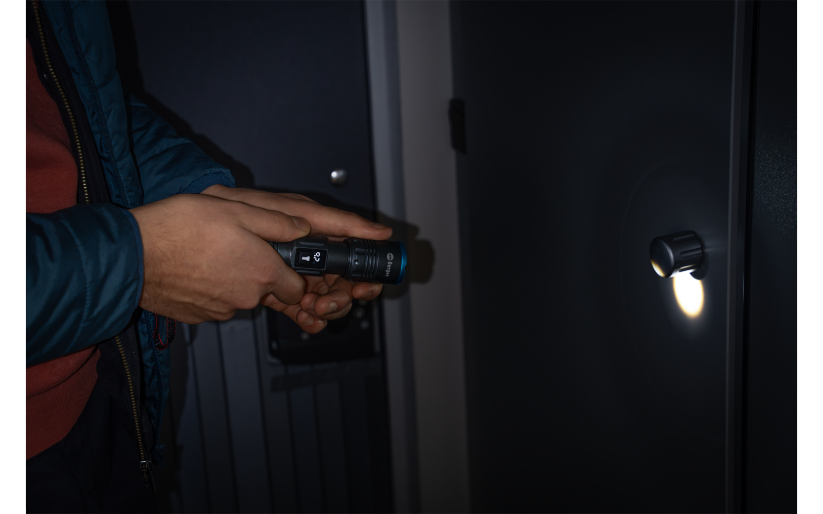 Hände einer Person in blauer Jacke und rotem Pullover halten eine Taschenlampe, deren Licht auf ein rundes Türschloss scheint