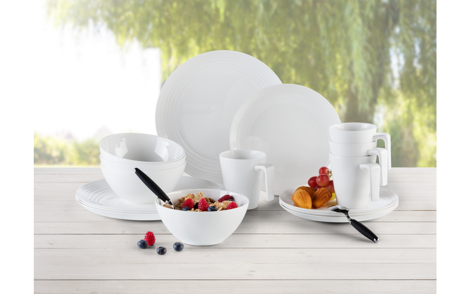 Weiße Teller, Schalen und Tassen auf einem hellen Tisch mit Obst und Besteck