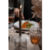 White plate with an orange‑brown elongated piece and green salad; a person cutting with knife and fork. Lit candle and two amber wine glasses on the table