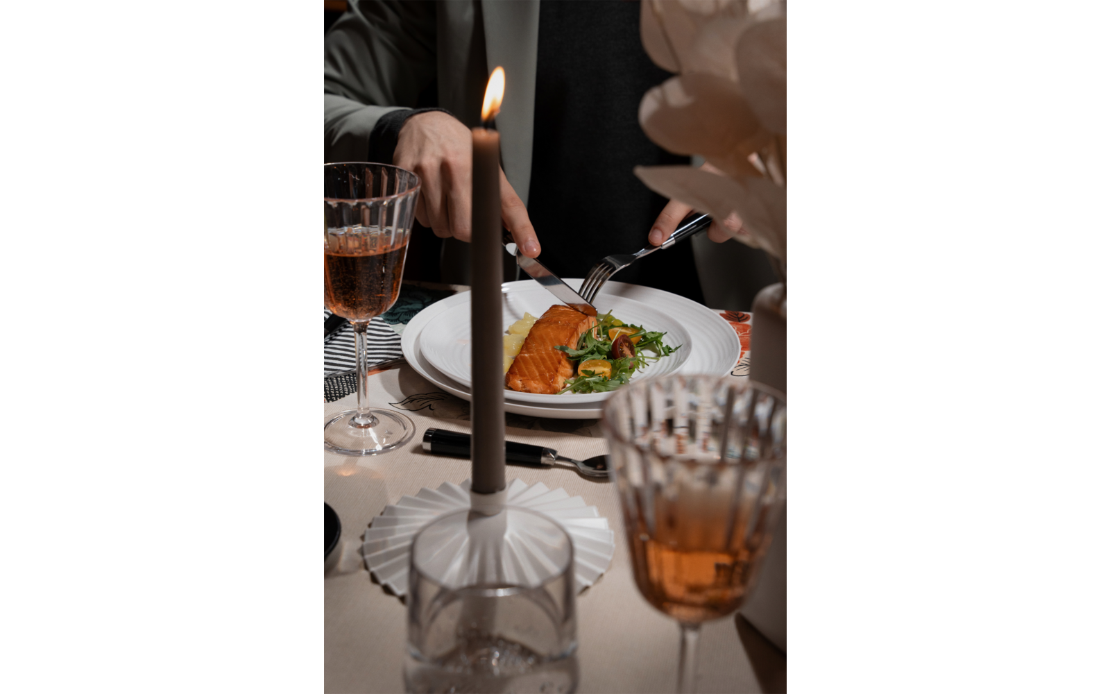 White plate with an orange‑brown elongated piece and green salad; a person cutting with knife and fork. Lit candle and two amber wine glasses on the table