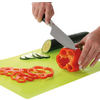 Two hands cutting a red bell pepper and a cucumber with a knife on a green cutting board; sliced pepper rings and cucumber slices beside them