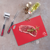 Raw steak on a red cutting board with a rosemary sprig, coarse salt, scattered peppercorns, two garlic cloves, glass bottle of oil with herbs, meat mallet and fork on a gray surface