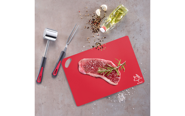 Raw steak on a red cutting board with a rosemary sprig, coarse salt, scattered peppercorns, two garlic cloves, glass bottle of oil with herbs, meat mallet and fork on a gray surface