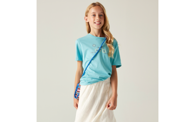 Mädchen mit langen blonden Haaren, hellblauem T‑Shirt, weißem Rock und bunter Umhängetasche mit blauem Riemen vor hellem Hintergrund