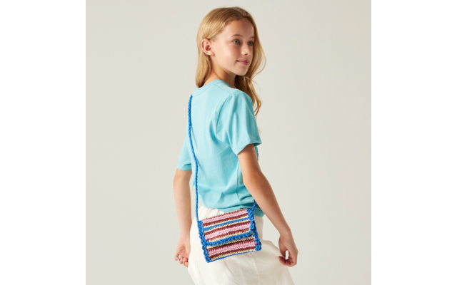 Seitlich stehendes Mädchen mit blondem Haar, türkisfarbenem T‑Shirt, weißem Rock und mehrfarbiger gehäkelter Umhängetasche mit blauem Riemen vor hellem Hintergrund
