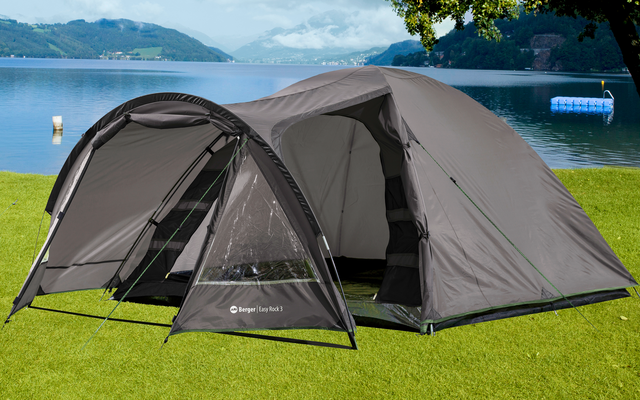 Zwei graue Zelte stehen auf einer grünen Wiese am Seeufer, im Hintergrund Berge und bewölkter Himmel