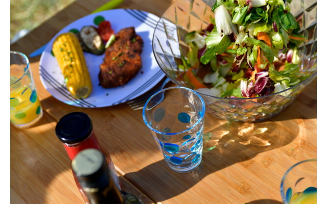 Wooden table with a glass bowl of salad, a blue plate with a corn on the cob, a piece of meat and vegetables, a clear glass with blue dots, and two bottles