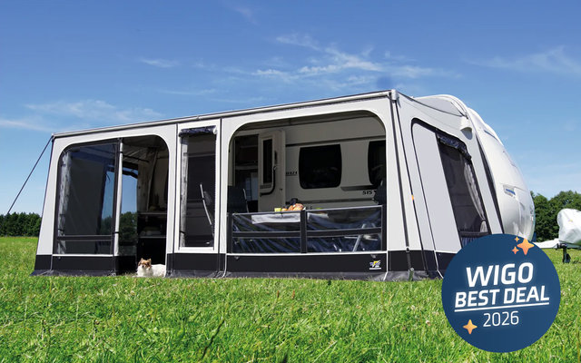 Wohnwagen mit Vorzelt auf einer Wiese am See; unter dem Vorzelt sitzt eine Person an einem Tisch, ein kleiner Hund liegt unter dem Tisch; im Hintergrund Wasser, Boote und Bäume
