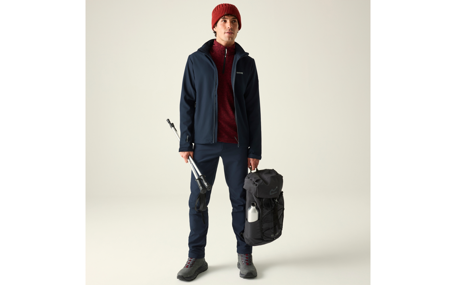 Person mit roter Mütze, rotem Reißverschluss-Pullover, dunkelblauer Jacke und Hose, hält ein zusammengeklapptes Dreibeinstativ in der rechten Hand und einen schwarzen Rucksack mit weißer Trinkflasche in der linken Hand, steht vor hellem Hintergrund