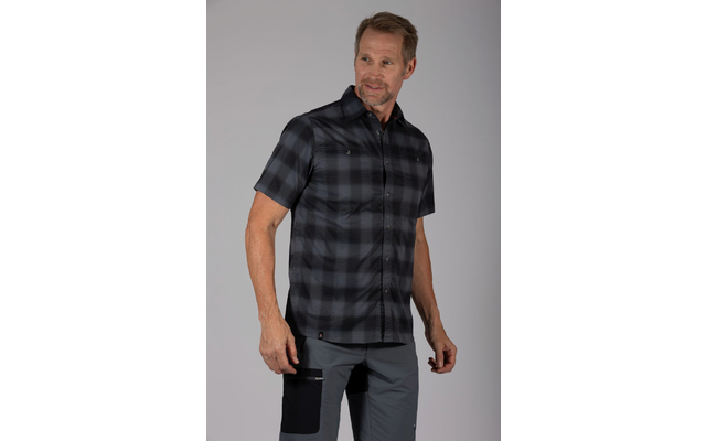 Man staat voor een effen lichtgrijze achtergrond, draagt een donker geruit kort mouw overhemd en grijze broek met een zwarte dijbeenzak, kijkt naar rechts, armen ontspannen langs zijn zij