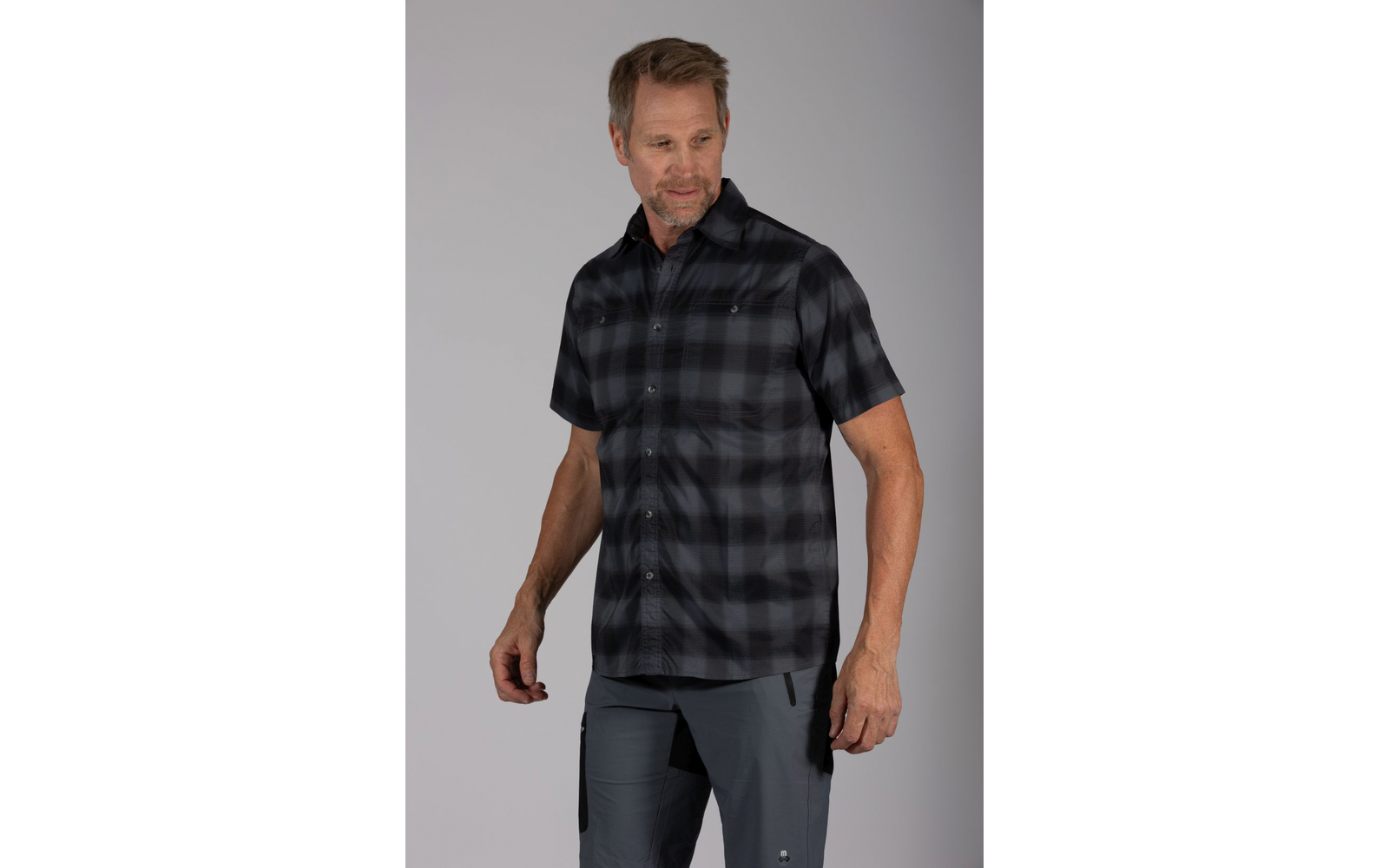 Een man staat voor een grijze achtergrond, draagt een donker geruit hemd met korte mouwen en een grijze broek, kijkt rechtsonder