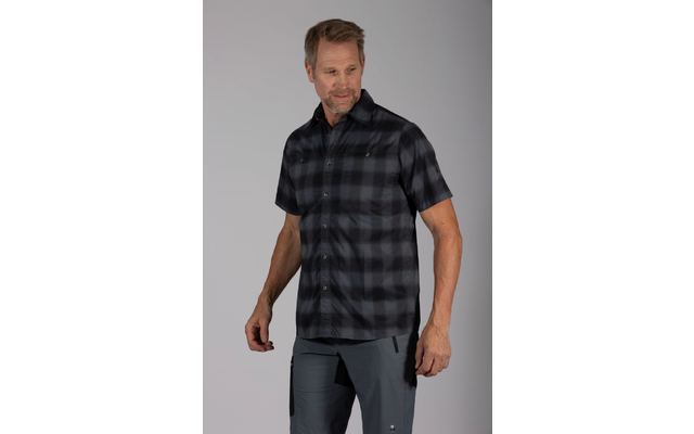 Een man staat voor een grijze achtergrond, draagt een donker geruit hemd met korte mouwen en een grijze broek, kijkt rechtsonder