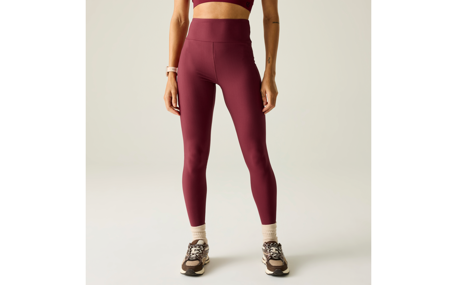 Vista parcial de un cuerpo de pie desde los hombros hasta los pies con mallas burdeos, un sujetador deportivo, calcetines claros, zapatillas deportivas y un reloj inteligente en la muñeca izquierda sobre fondo claro