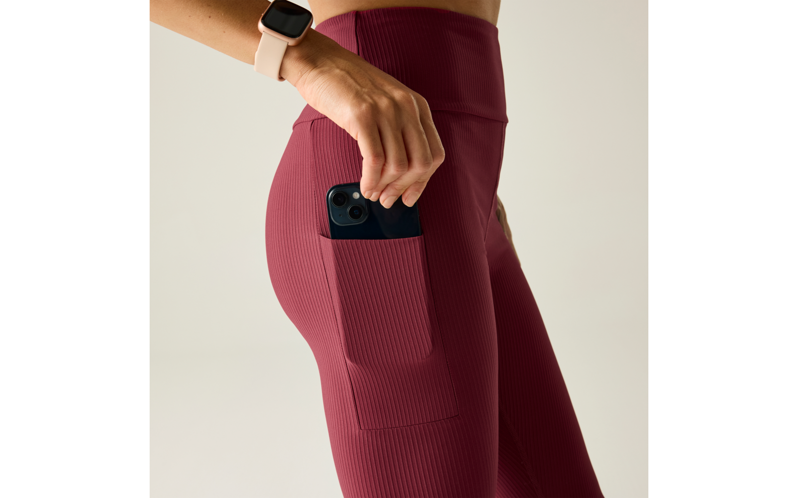 Primer plano de una persona que coloca un teléfono inteligente oscuro en el bolsillo lateral de unas mallas burdeos con canalé; mano con reloj inteligente claro visible