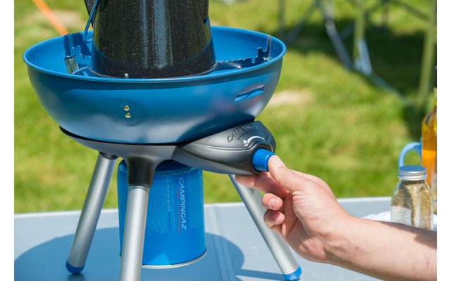 Hand drukt blauwe knop op draagbare blauwe campinggaskooker met gasbus en drie poten, op een tafel; gras op de achtergrond
