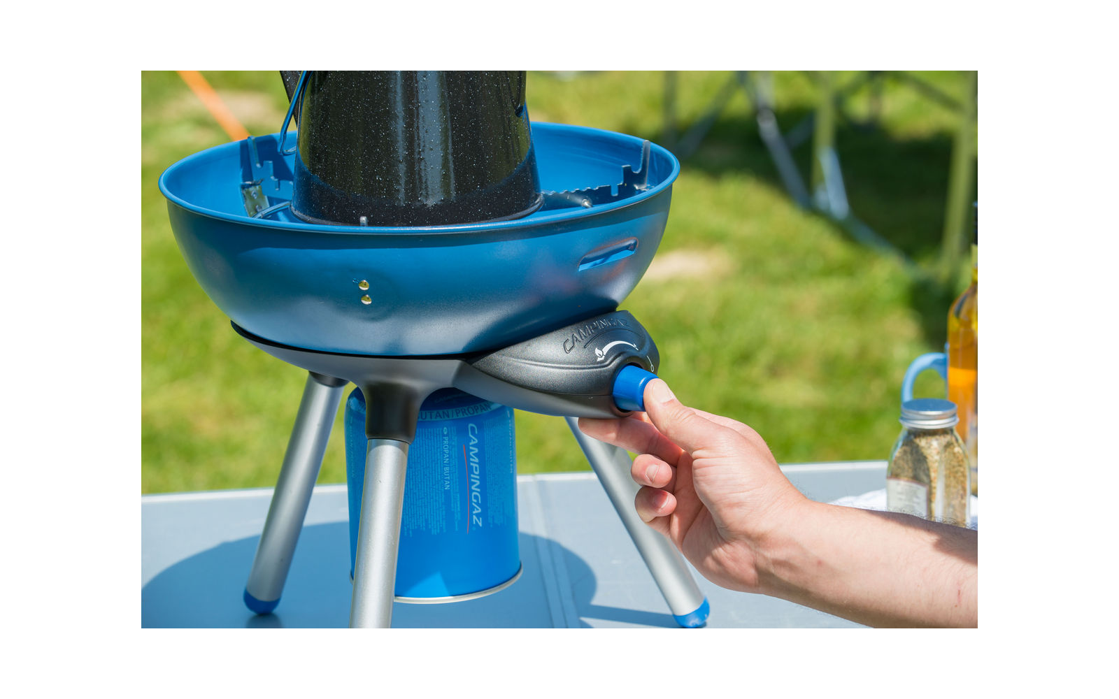 Hand drukt blauwe knop op draagbare blauwe campinggaskooker met gasbus en drie poten, op een tafel; gras op de achtergrond