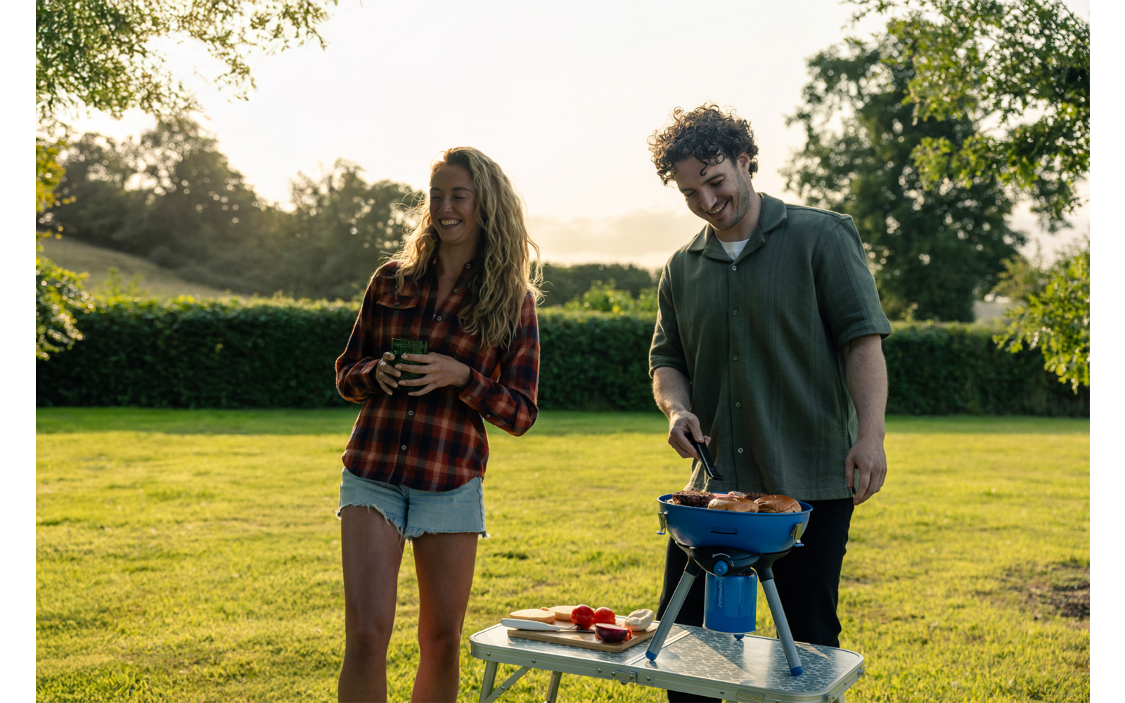 Twee personen op een grasveld; vrouw in geruit shirt en korte spijkerbroek houdt een beker vast, man in groen shirt bedient een kleine blauwe barbecue op een tafel; tomaten op de tafel; bomen en haag op de achtergrond