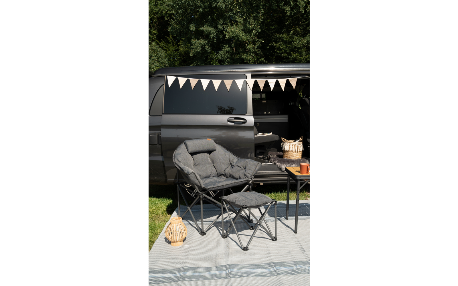 Campingstuhl mit Fußbank vor einem geöffneten Bulli mit Girlande, daneben ein kleiner Tisch mit Korb und Becher