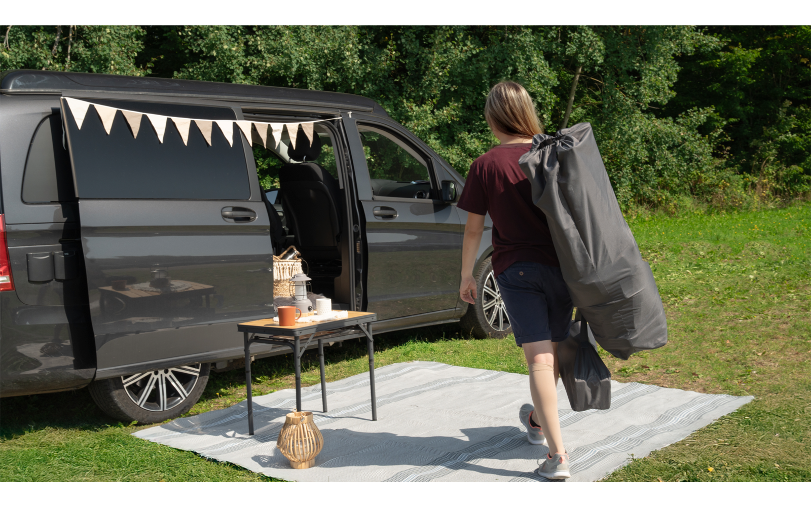 Person trägt eine große graue Tasche und geht auf einen schwarzen Van mit offener Schiebetür zu, davor steht ein Tisch auf einer Matte im Gras