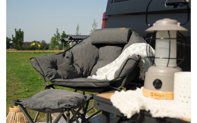 Grauer gepolsterter Sessel mit Decke und Hocker im Garten, daneben eine Laterne auf einem Tisch