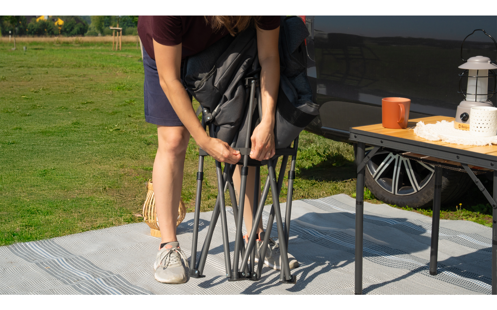 Person faltet einen tragbaren Stuhl auf einer Decke neben einem Tisch mit Becher und Laterne