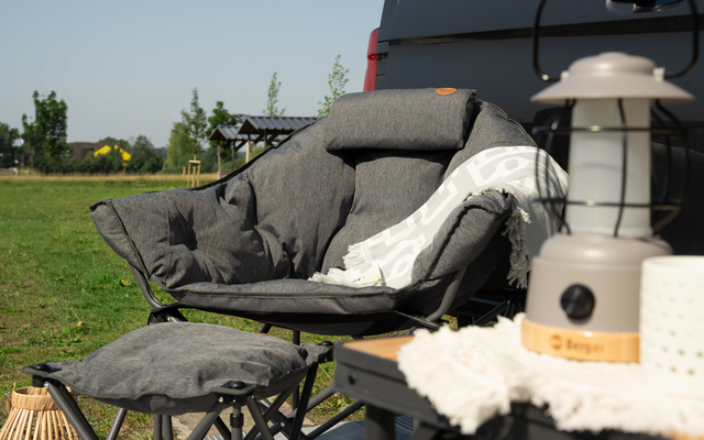 Fauteuil rembourré gris avec couverture et repose-pieds dans le jardin, à côté d'une lanterne sur une table