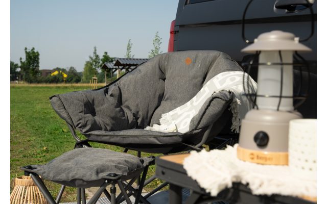 Gray padded armchair with blanket and footstool in the garden, next to a lantern on a table