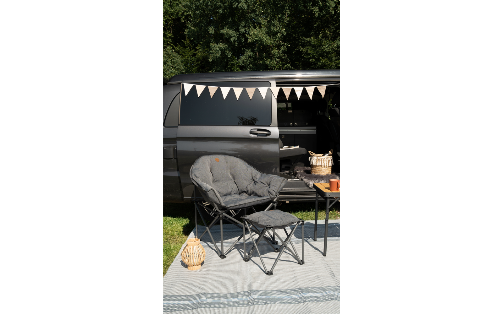 Camping chair with footrest in front of an open camper van decorated with a garland, next to a small table with a basket and cup
